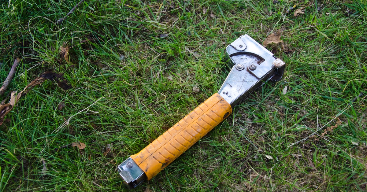 A well-used yellow and silver hammer tacker sitting on the grass, surrounded by brown leaves and twigs.