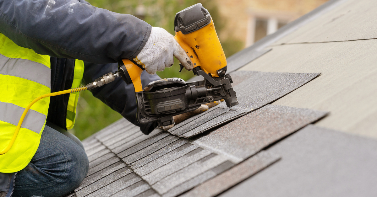 A construction worker kneels on a gray roof using a pneumatic nail gun to staple a tile. They wear gloves while using the tool.