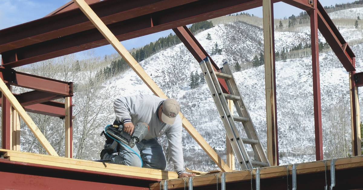 A person using a nail gun on a wooden platform with steel beams in the foreground and snowy mountains behind.