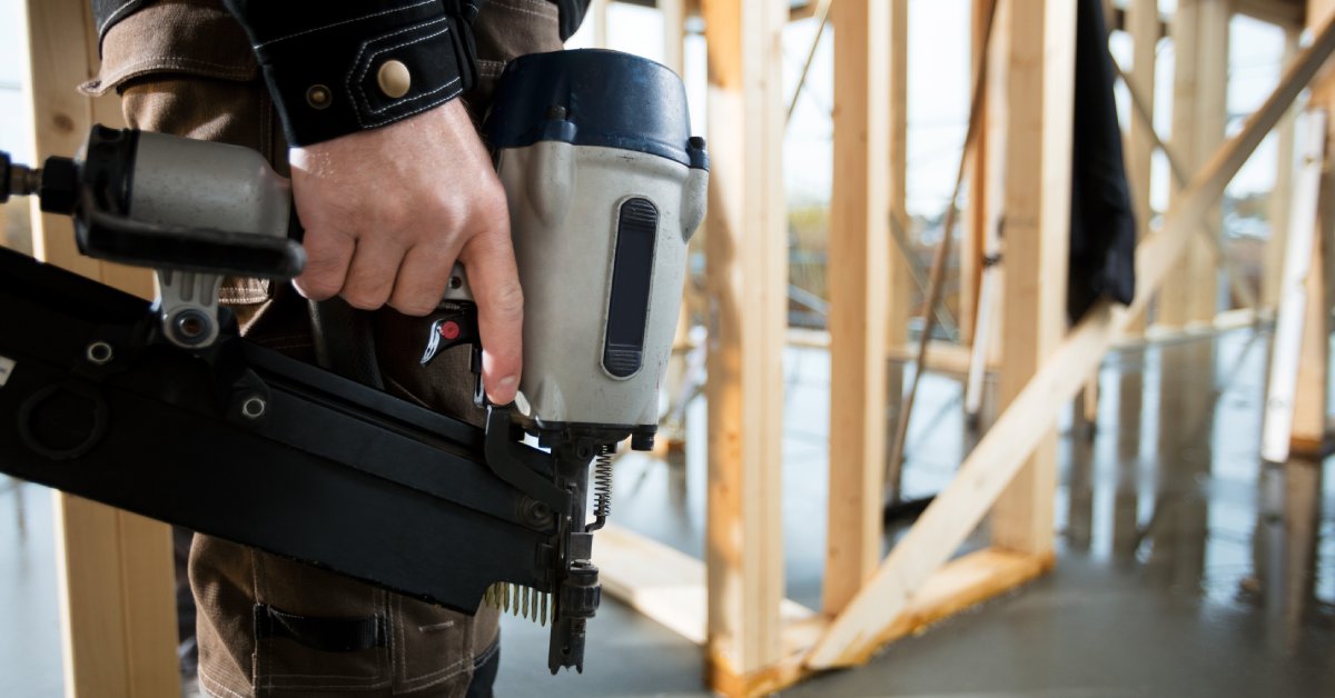 A man wearing brown pants and a blue shirt stands in an unfinished home while holding a finish nailer nailgun.