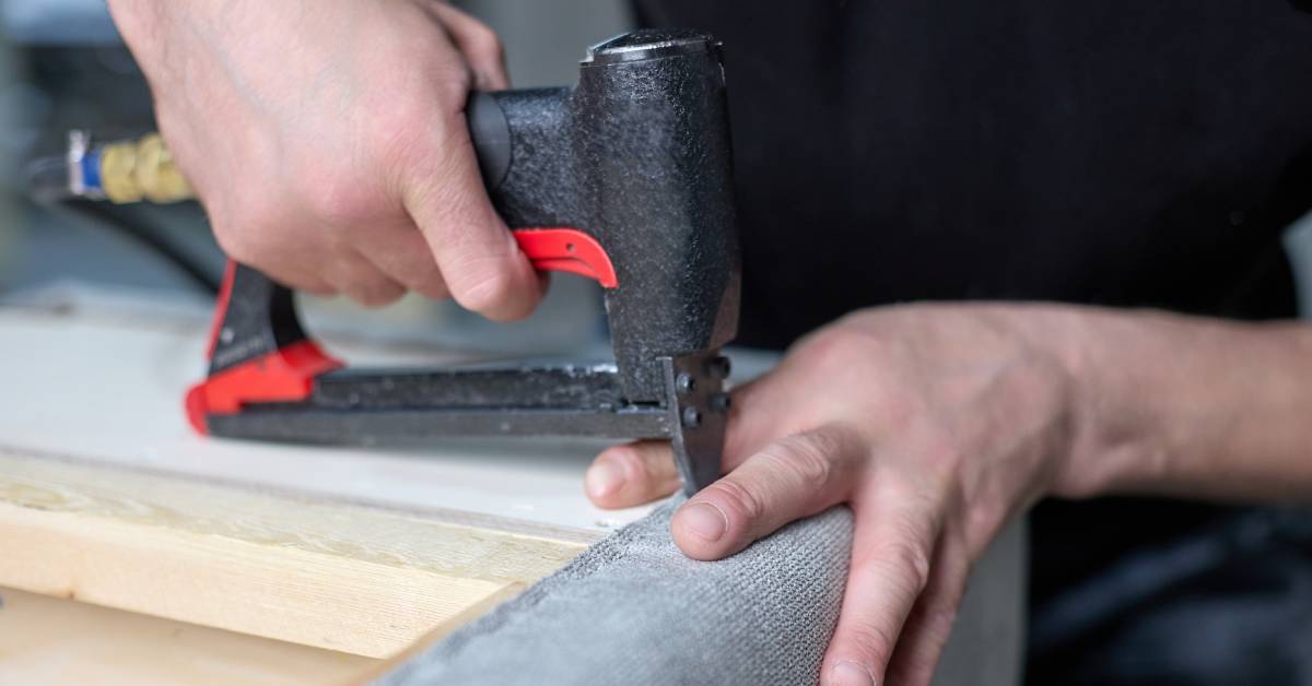 A person using a pneumatic staple gun to attach the fabric to a wooden surface in an upholstery project.