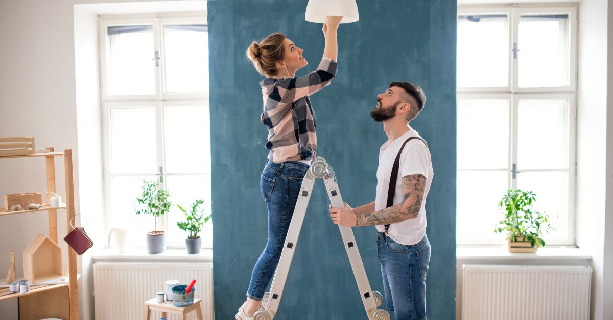 A man holds a ladder steady as a woman changes a light fixture in a bright room with plants and paint supplies nearby.