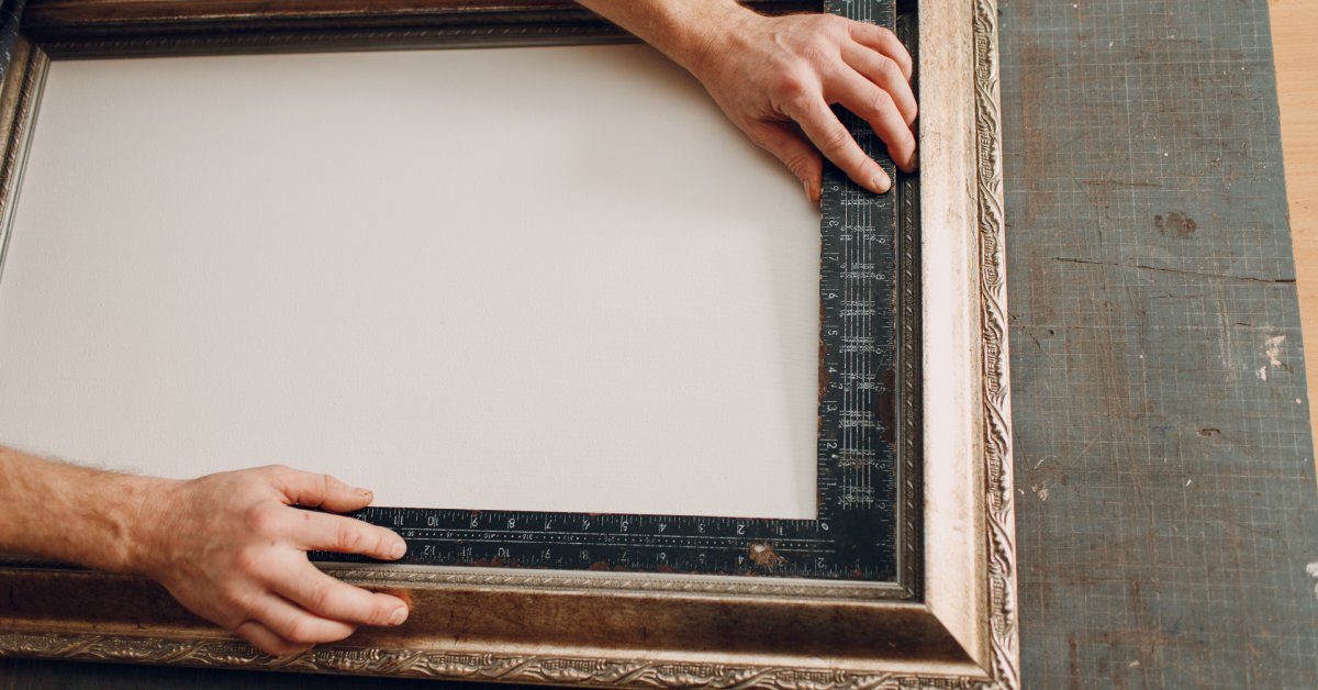 A person's hand uses a ruler to measure the corners of a bronze, baguette picture frame with white backing.