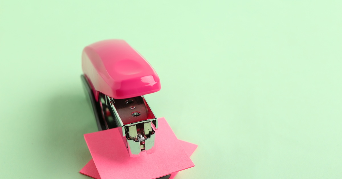 A pink stapler has a stack of pink sticky notes placed beneath the hammer. The background is mint green.
