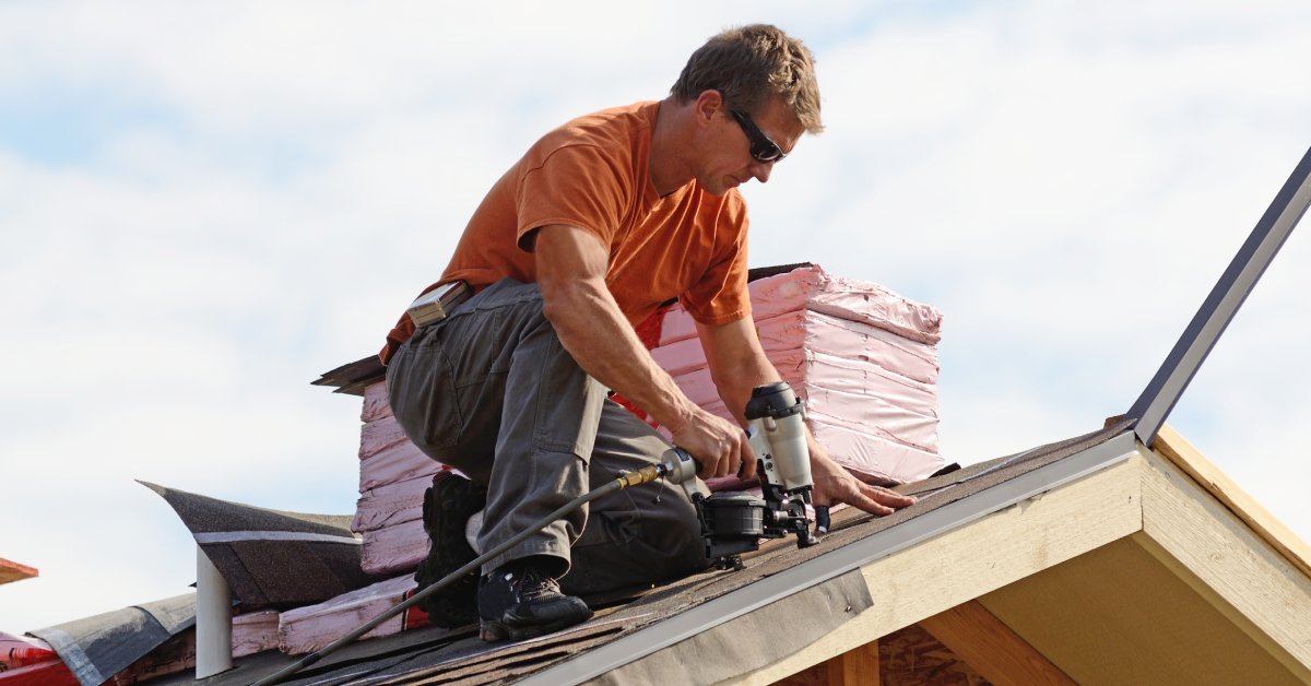 A man in an orange shirt uses a nail gun on a rooftop under construction, with pink insulation in the background.