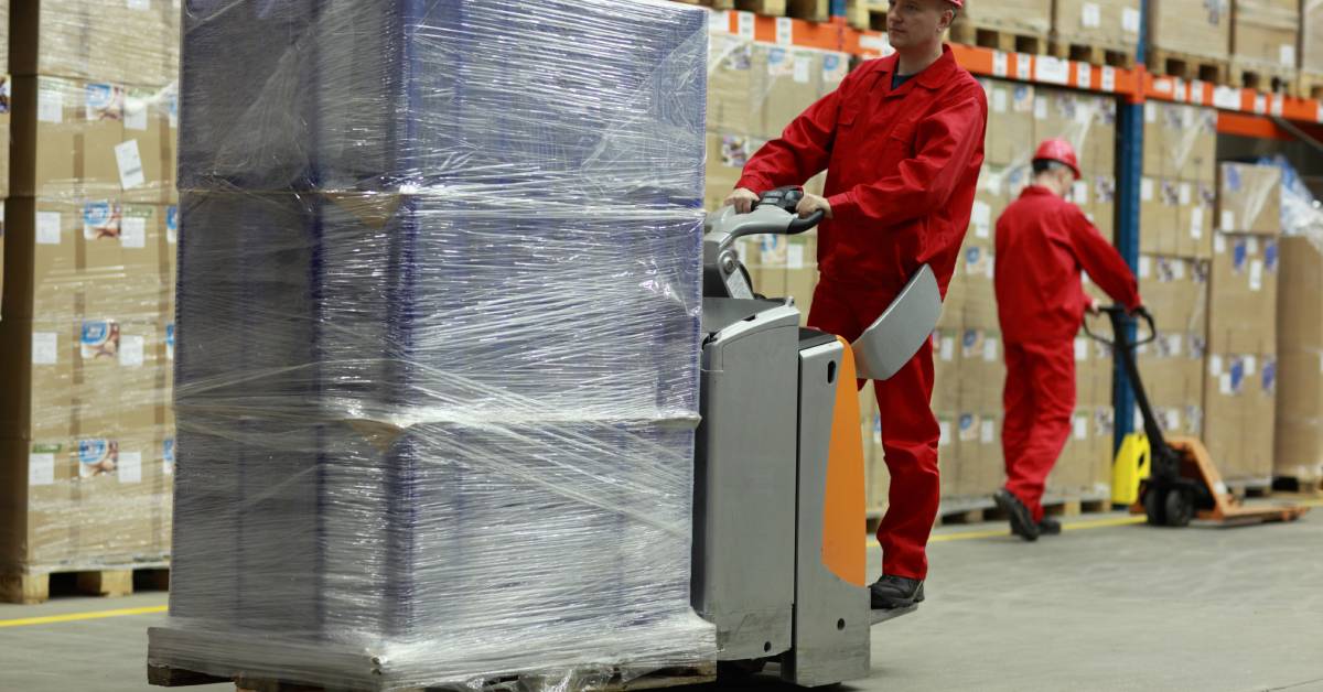 Two workers in red uniforms and helmets operate pallet jacks in an organized warehouse filled with stacked boxes on shelves.