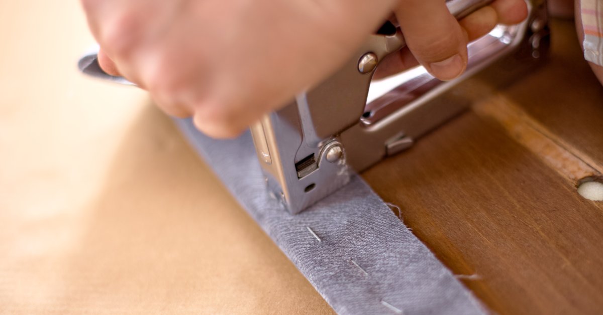 A close-up of a hand pressing a silver staple gun onto gray fabric stretched over a wooden surface, securing it with staples.
