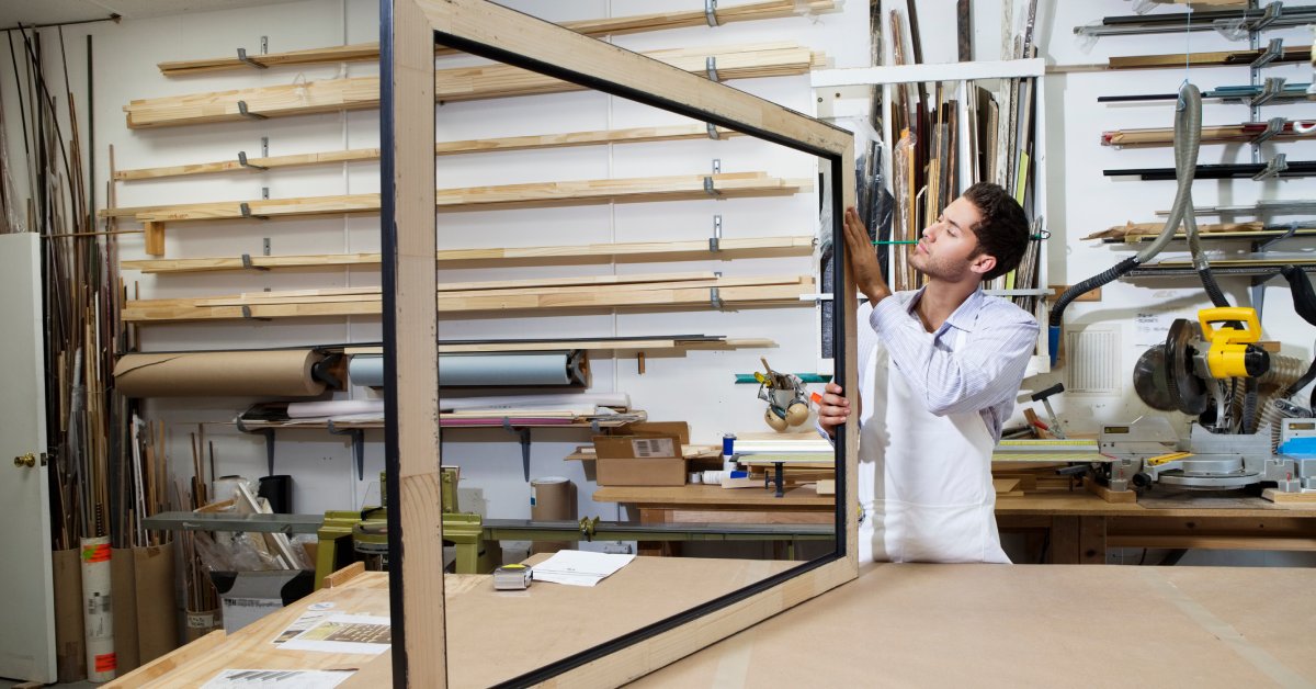 A person in a white apron holds a large wooden frame in a workshop with shelves, tools, and materials in the background.