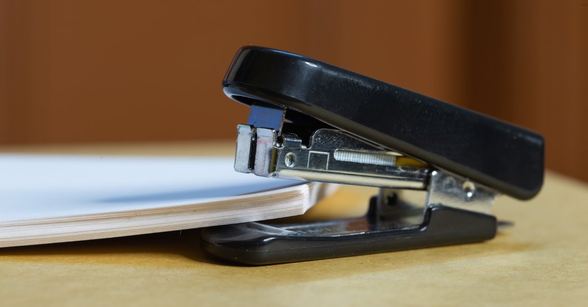 A stack of paper rests in the mouth of a black metal stapler on a wooden surface. The edges of the stapler are round.