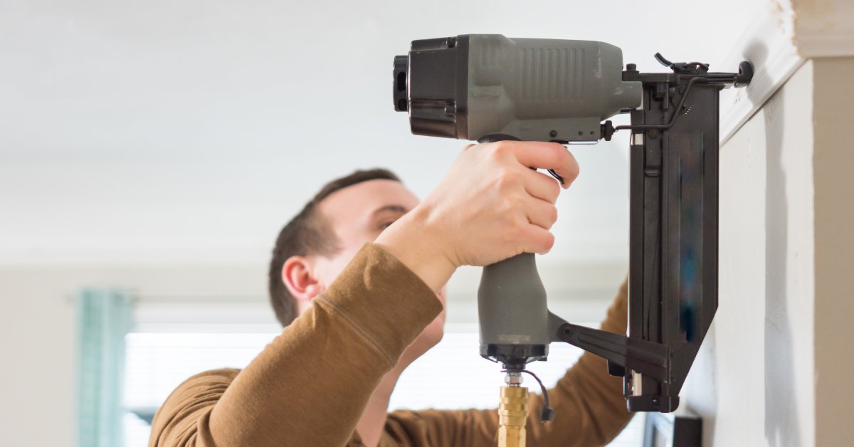 A Caucasian man wearing a brown sweater uses a gray brad nail gun to attach a white trim on a beige wall.