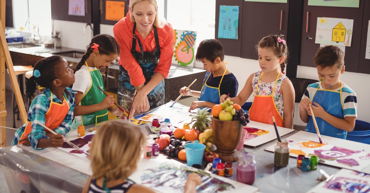A teacher in a red shirt and apron stands by children painting on canvases. The table has paint, cups, and paintbrushes.