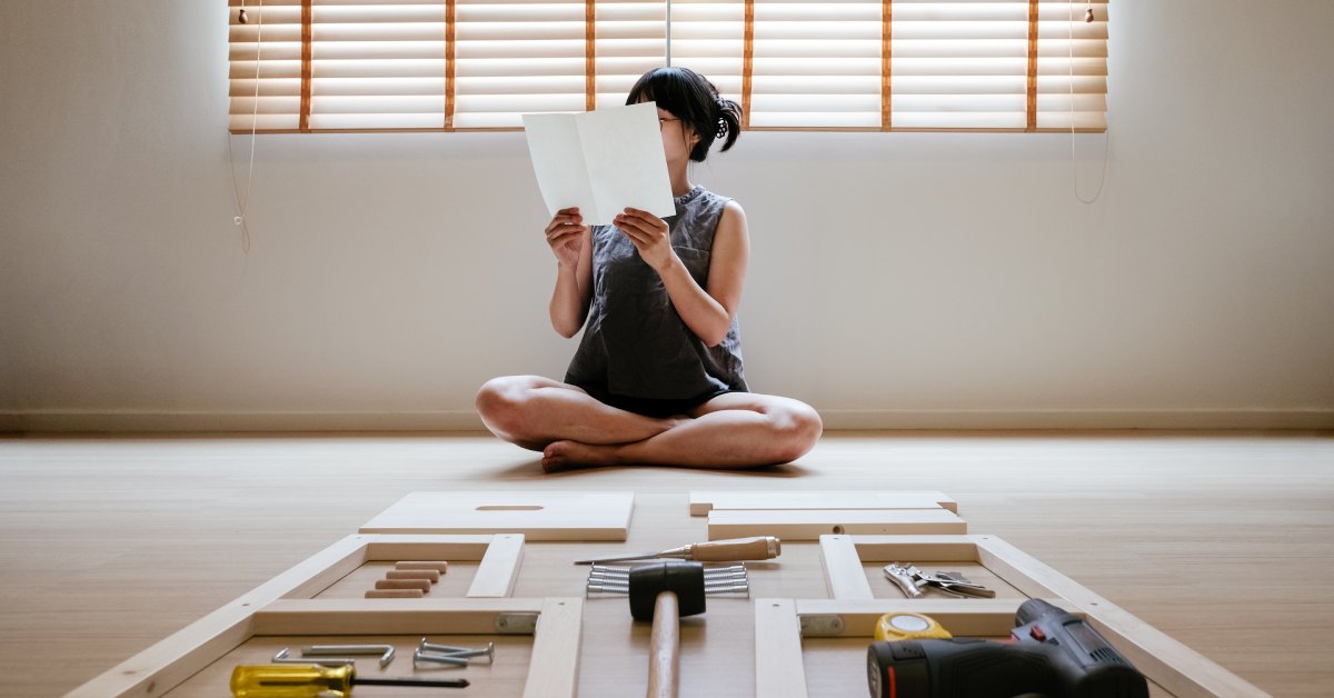 A woman sits cross-legged on the floor, surrounded by wood blocks and tools, studying a piece of paper.