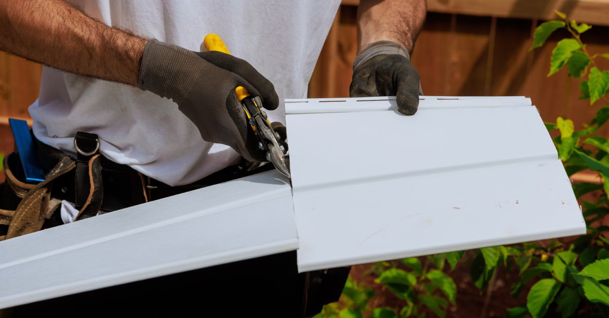 A person wearing gloves uses tin snips to cut a piece of white vinyl siding, with plants and a wooden fence nearby.
