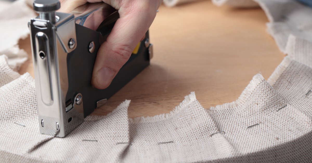 A person's hand using a black manual stapler to secure sturdy, light-colored fabric to wooden furniture.