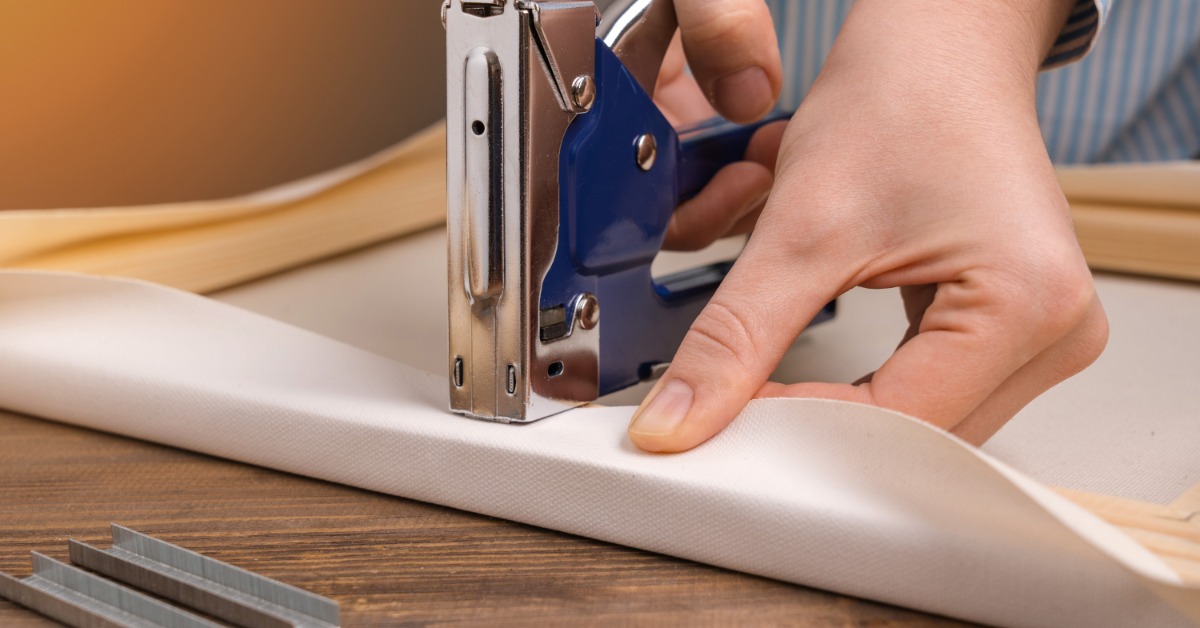 A close-up of an artist's hands stapling a canvas onto a stretched-out wooden frame in a studio workspace.