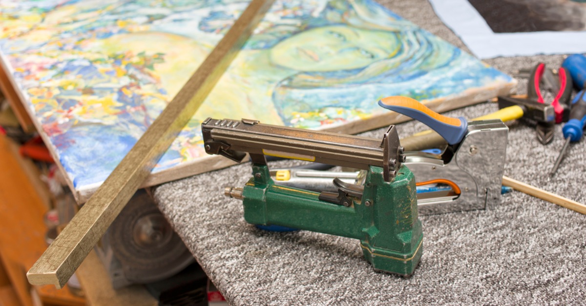 A studio table with a stapler, a ruler, and frame moulding on top, as a framer assembles a handmade picture frame.