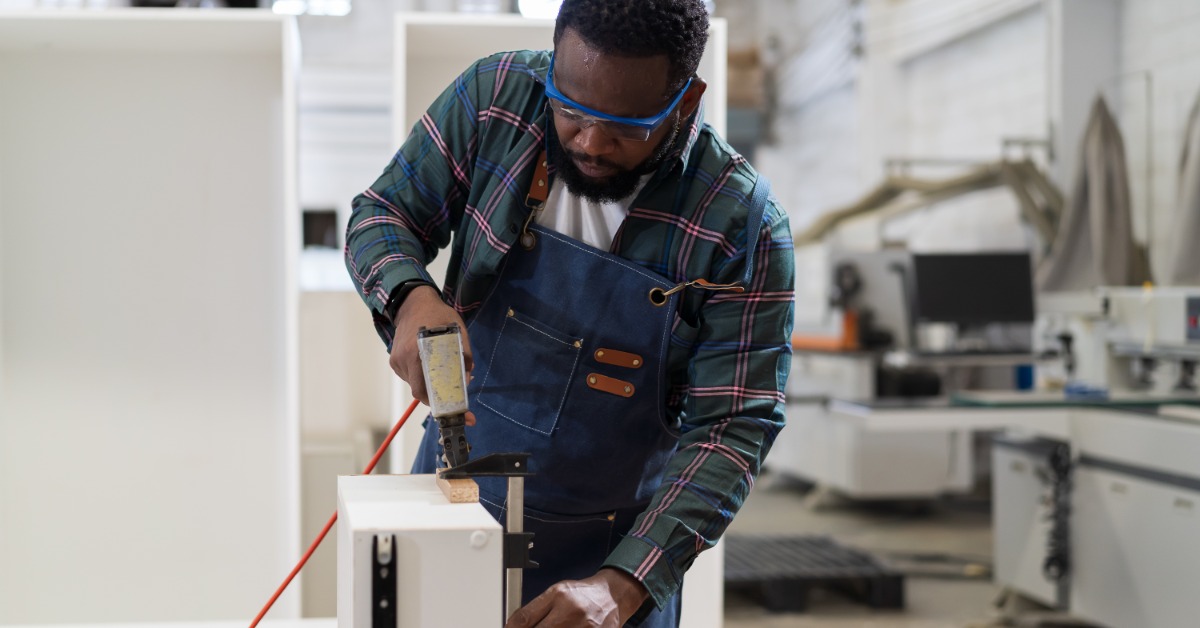 Carpenter assembling furniture uses a stapler to attach fabric to a wooden frame on a workbench in a workshop.