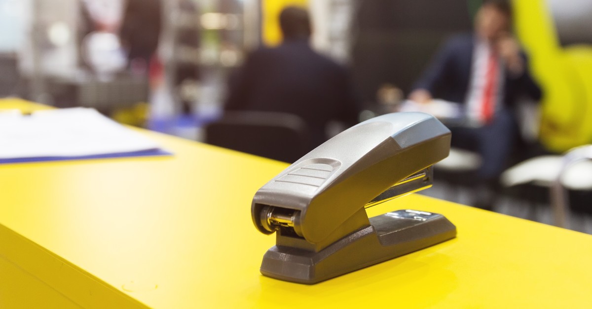 Overhead light reflects on a black stapler sitting on a yellow table, with a group of people in the background.