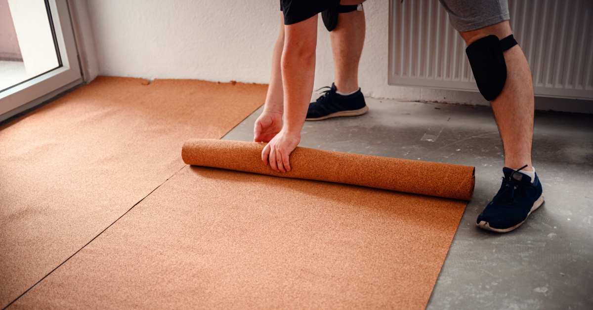 A person wearing gray shorts and black running shoes rolls out a brown underlayment on the cement floor.