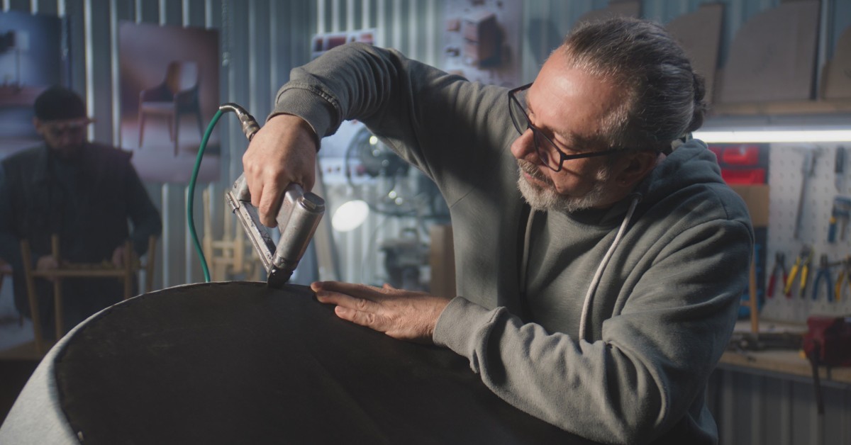 An older male wearing glasses and a gray sweater uses a staple gun to attach the fabric on a piece of furniture.